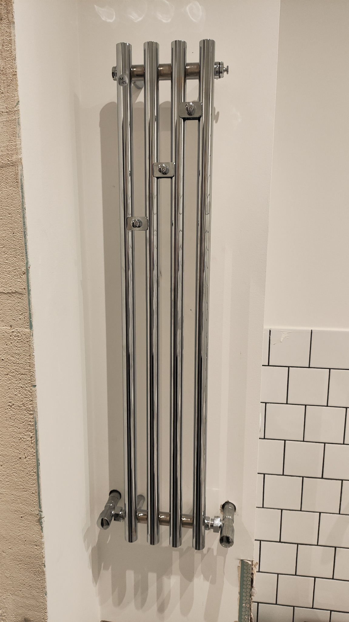 Błyszczący, chromowany grzejnik łazienkowy drabinkowy, zamontowany na białej ścianie, z widocznymi przyłączami hydraulicznymi i fragmentem ściany z białymi płytkami.