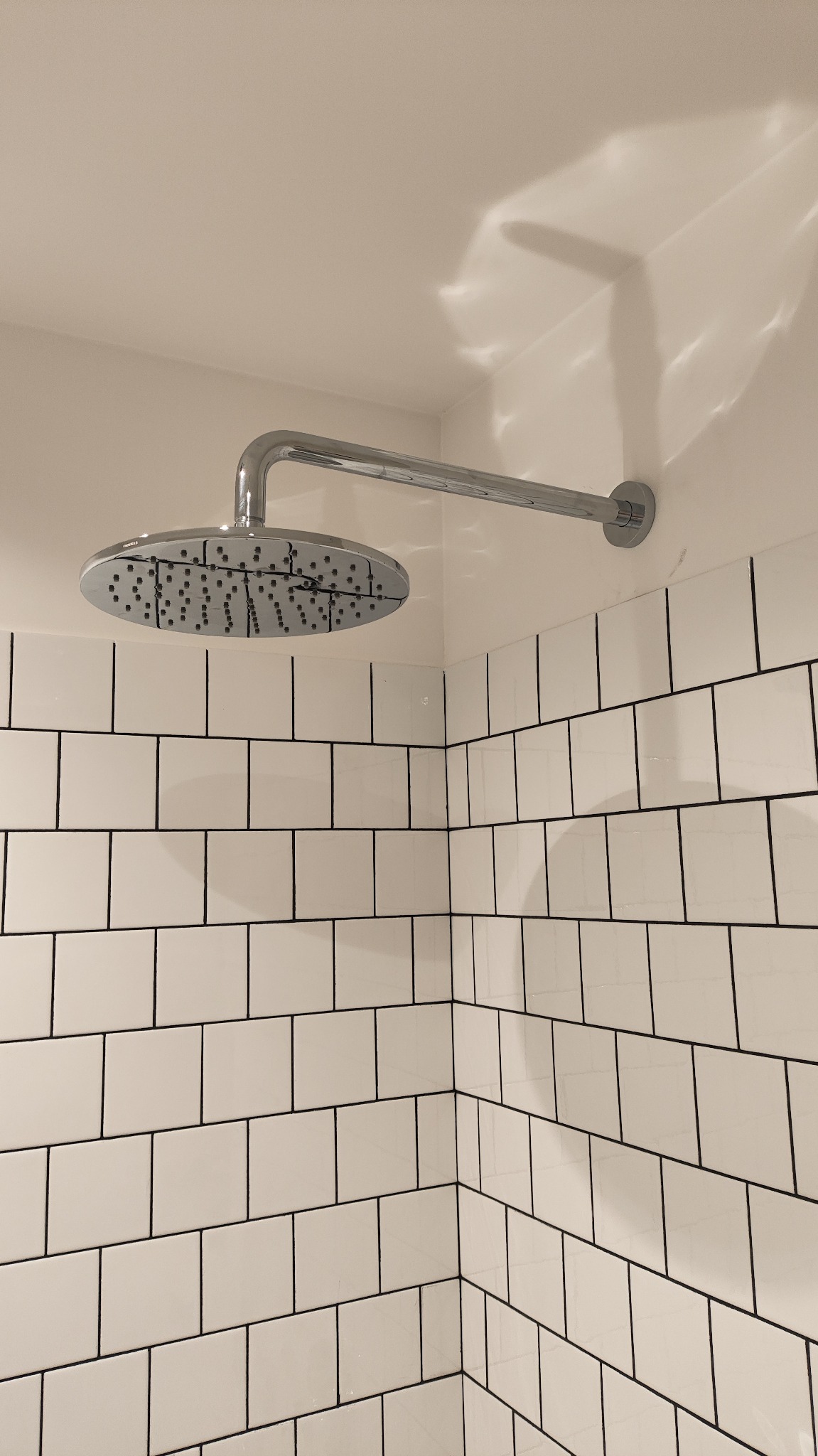 Nowoczesna, chromowana deszczownica zamontowana na białej ścianie z płytek typu metro z czarną fugą. Minimalistyczny design, oświetlenie sufitowe.