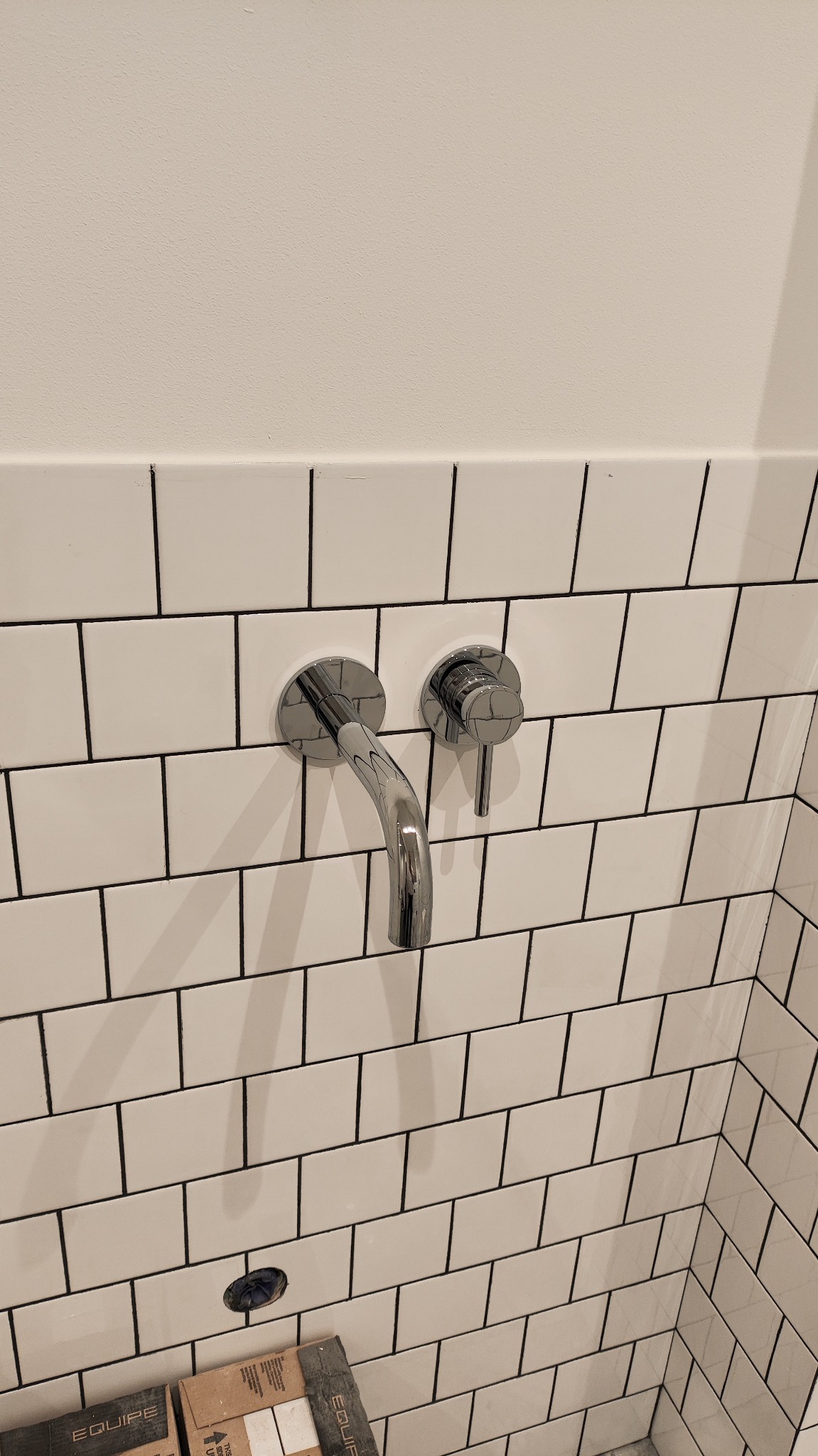 Bateria umywalkowa chromowana, zamontowana na ścianie z białych płytek z czarną fugą. Widoczne pudełka po płytkach. Minimalistyczny design.