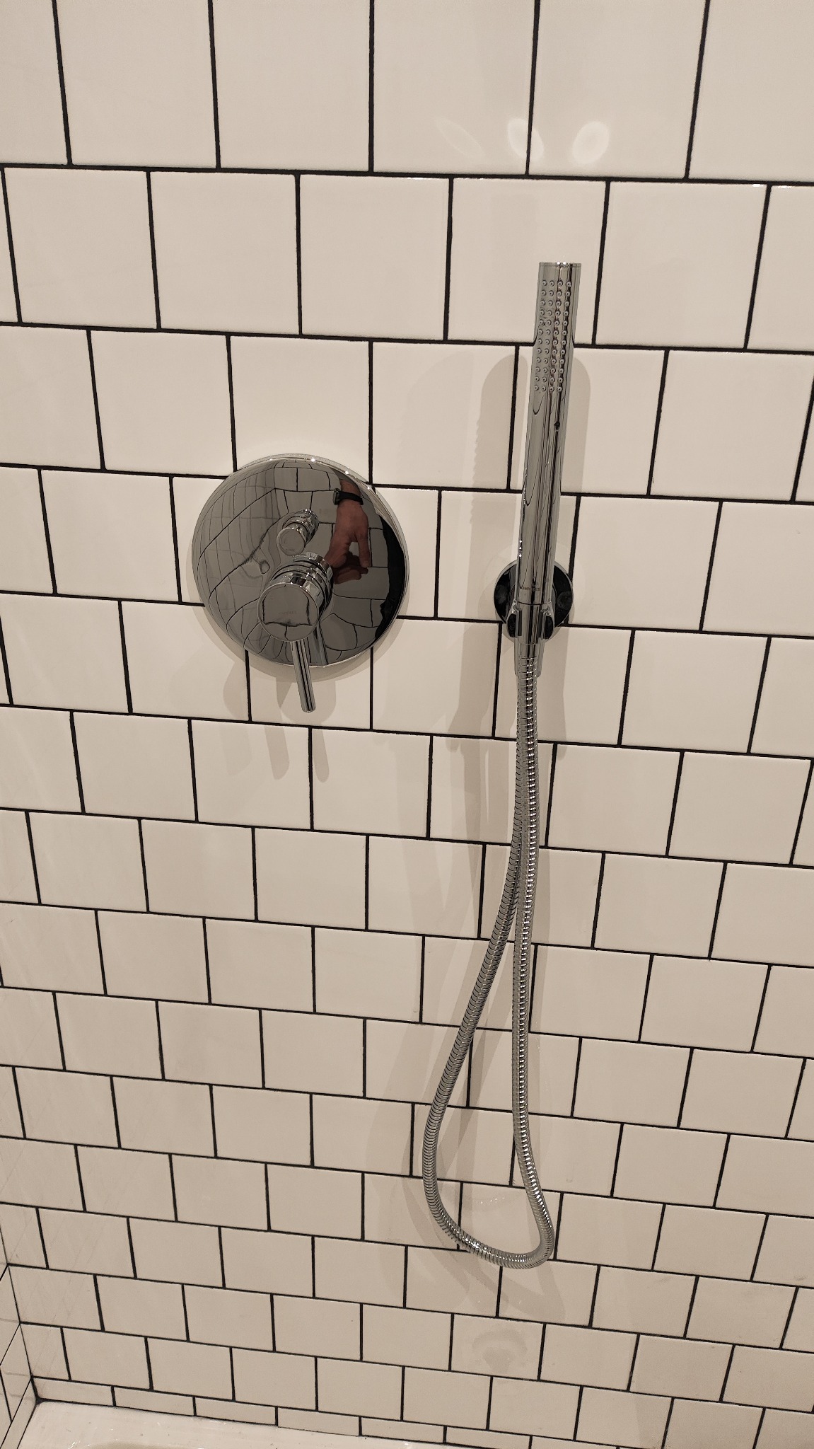 Bateria prysznicowa z deszczownicą na białej ścianie z czarną fugą. Chromowana armatura łazienkowa, widoczne odbicie dłoni na baterii.