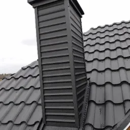 Czarny, blaszany komin wentylacyjny na dachu z czarnej blachodachówki, widoczne narzędzia dekarskie na szczycie komina.