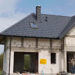 Dom w stanie surowym zamkniętym w Płońsku z nowym, grafitowym dachem z oknem dachowym i kominem wentylacyjnym. Widoczne orynnowanie i fragment elewacji z pustaków.