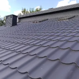 Nowoczesne krycie dachu z blachy trapezowej w kolorze grafitowym, z widocznym kominem i obróbkami blacharskimi, na tle błękitnego nieba.