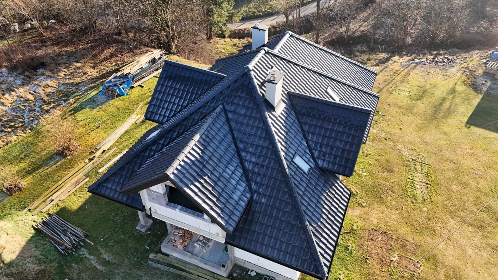 Widok z lotu ptaka na dom w trakcie budowy, z nowym, ciemnym dachem z dachówki ceramicznej, kominy wykończone na biało, widoczne składowane materiały budowlane na trawniku.