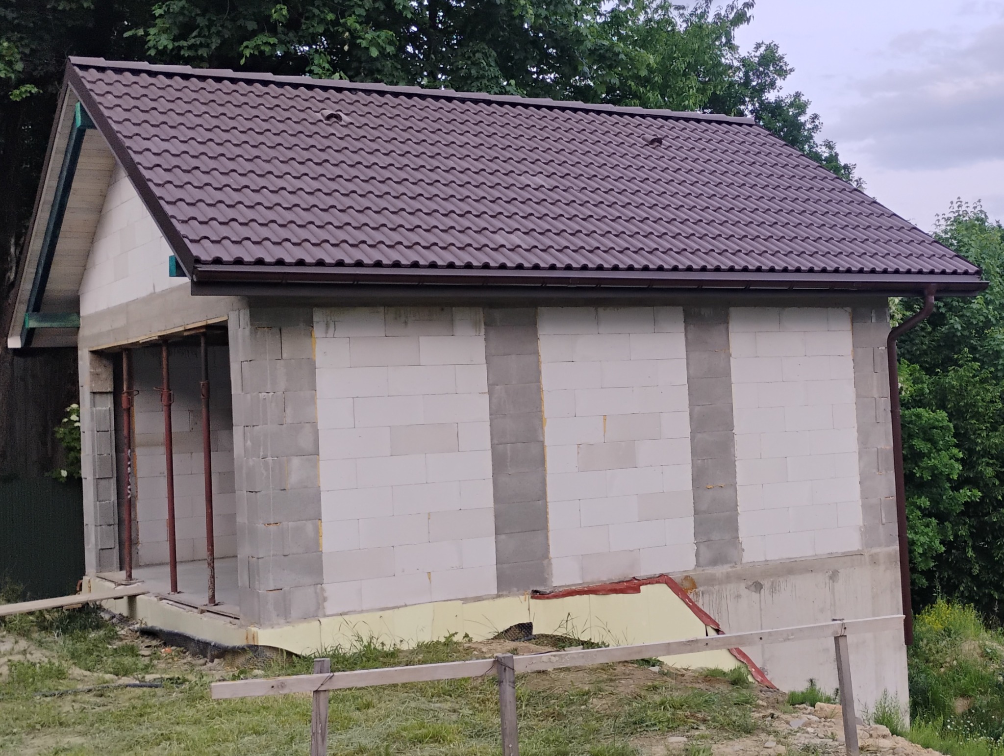 Niedokończony budynek z brązowym dachem z blachodachówki, widoczne białe ściany z betonu komórkowego, podparte metalowymi stemplami i fragment zieleni.