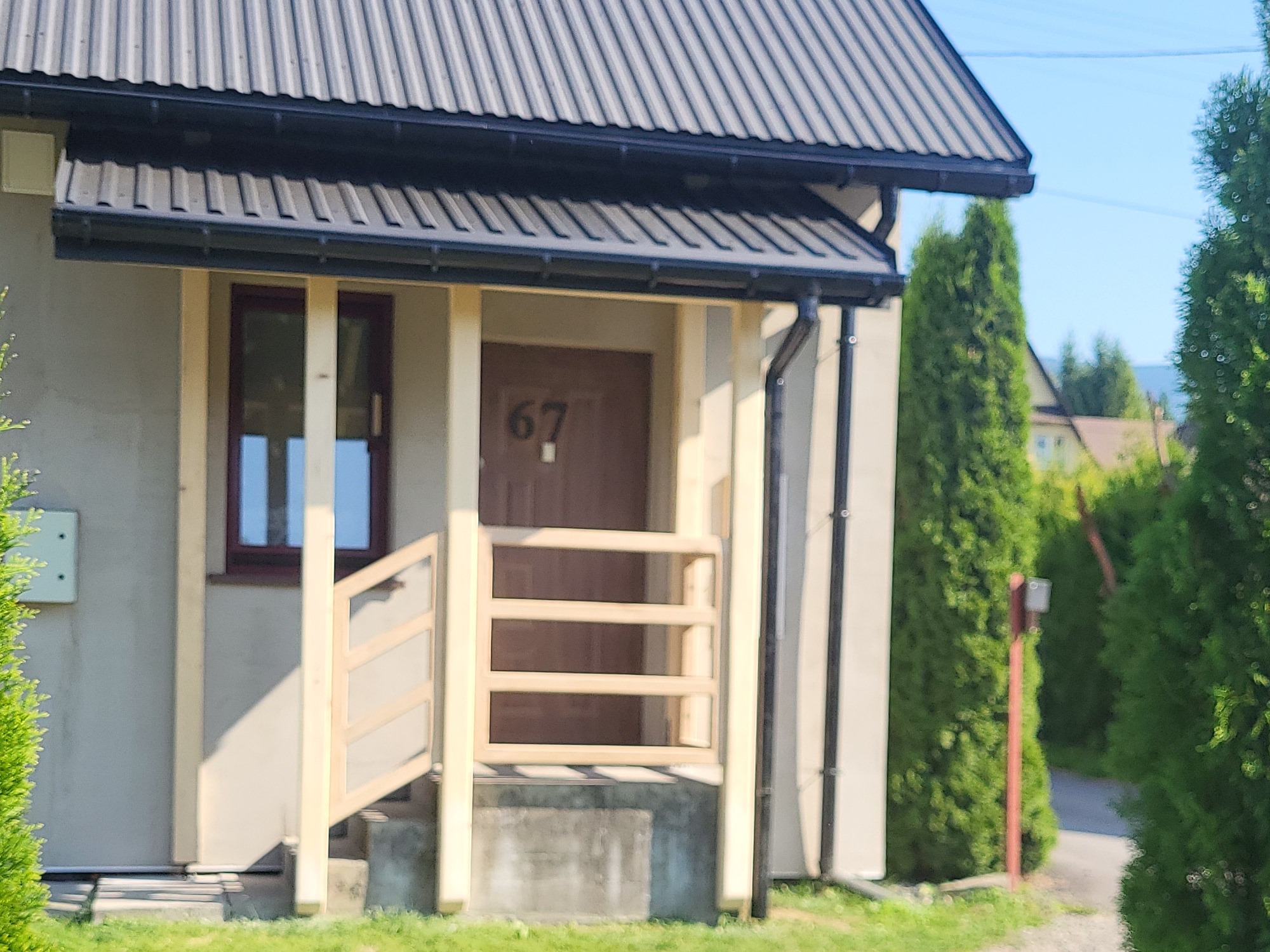 Wejście do domu z numerem 67, zadaszone czarnym dachem z blachy trapezowej, jasna elewacja, drewniana balustrada i okno z brązową ramą. W tle zieleń.