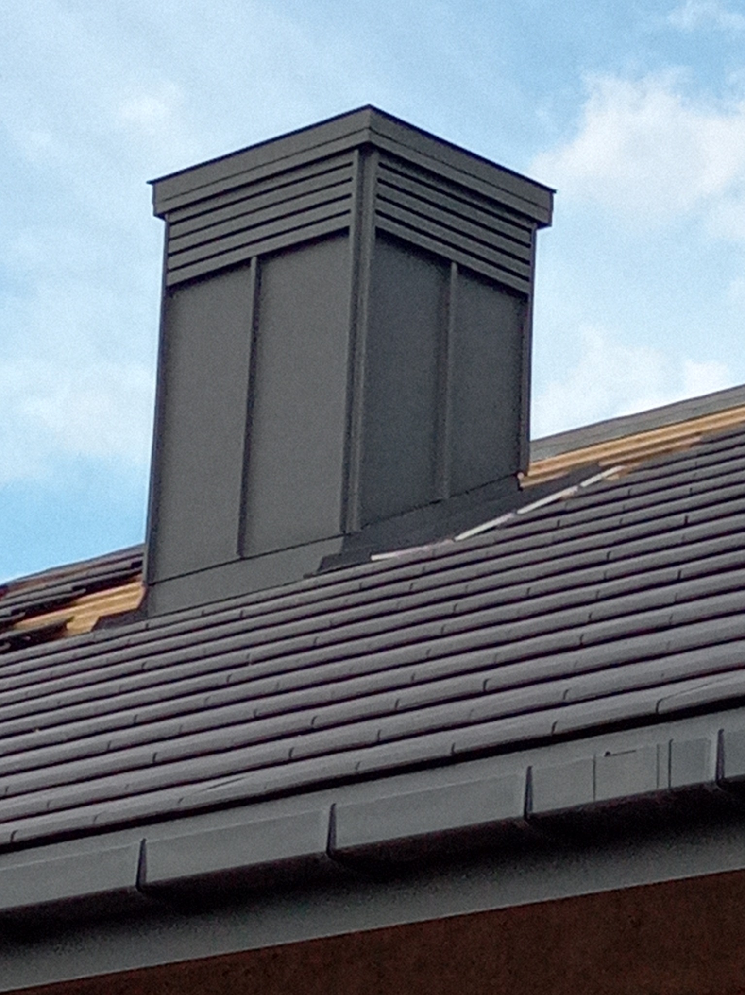 Grafitowy komin wentylacyjny na dachu z dachówki ceramicznej w Juszczynie. Widoczne elementy konstrukcji dachu. Ujęcie z dołu, na tle błękitnego nieba.