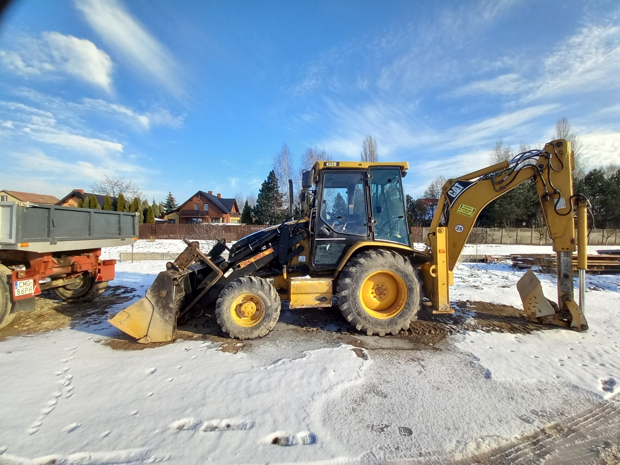 Żółto-czarna koparko-ładowarka Caterpillar 432D na ośnieżonym terenie, z widoczną przyczepą i domami w tle pod błękitnym niebem z chmurami.