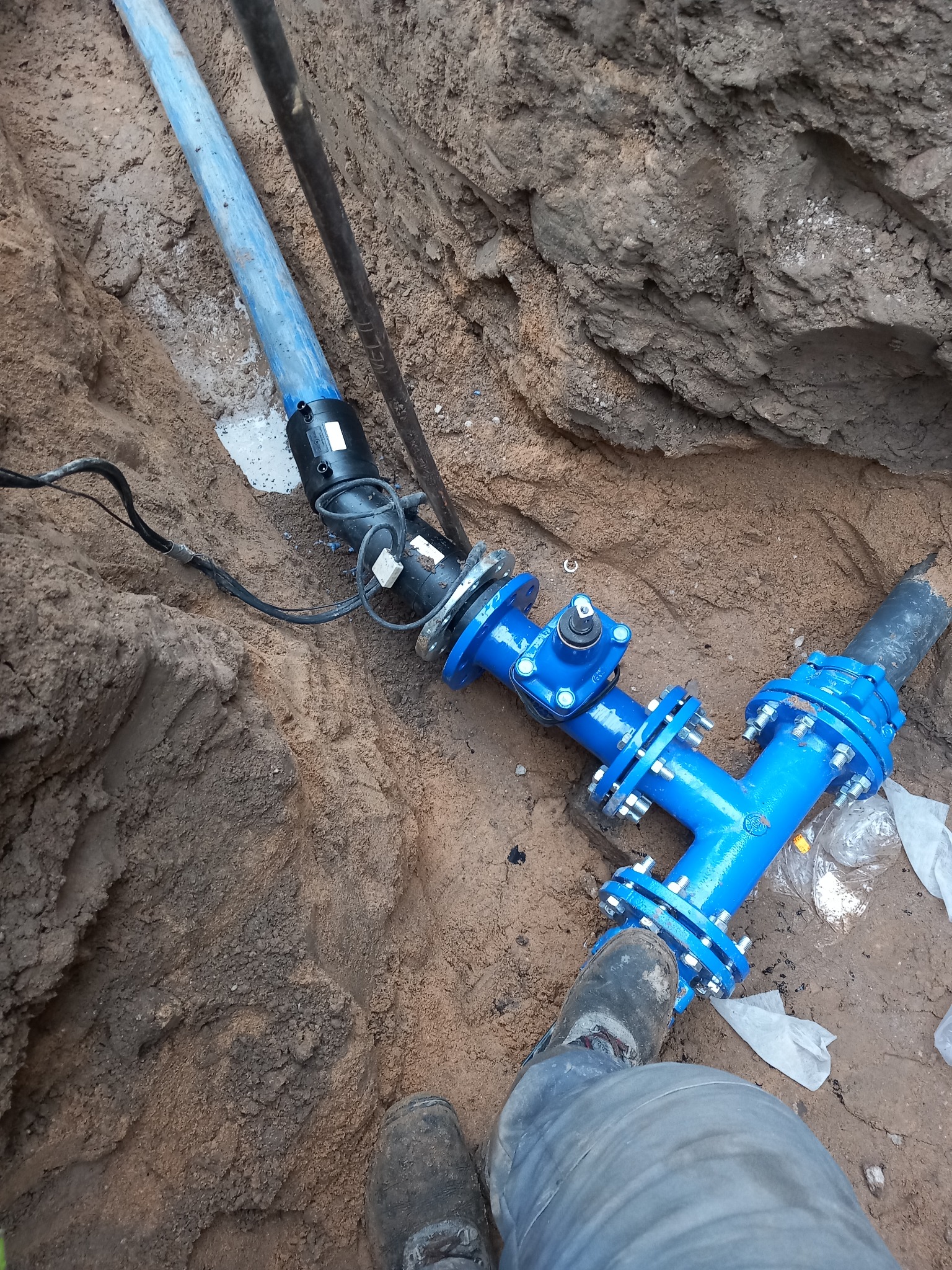 Niebieski trójnik z kołnierzami i śrubami, część podziemnej instalacji wodociągowej, widoczny w wykopie ziemnym z fragmentem buta roboczego w kadrze.