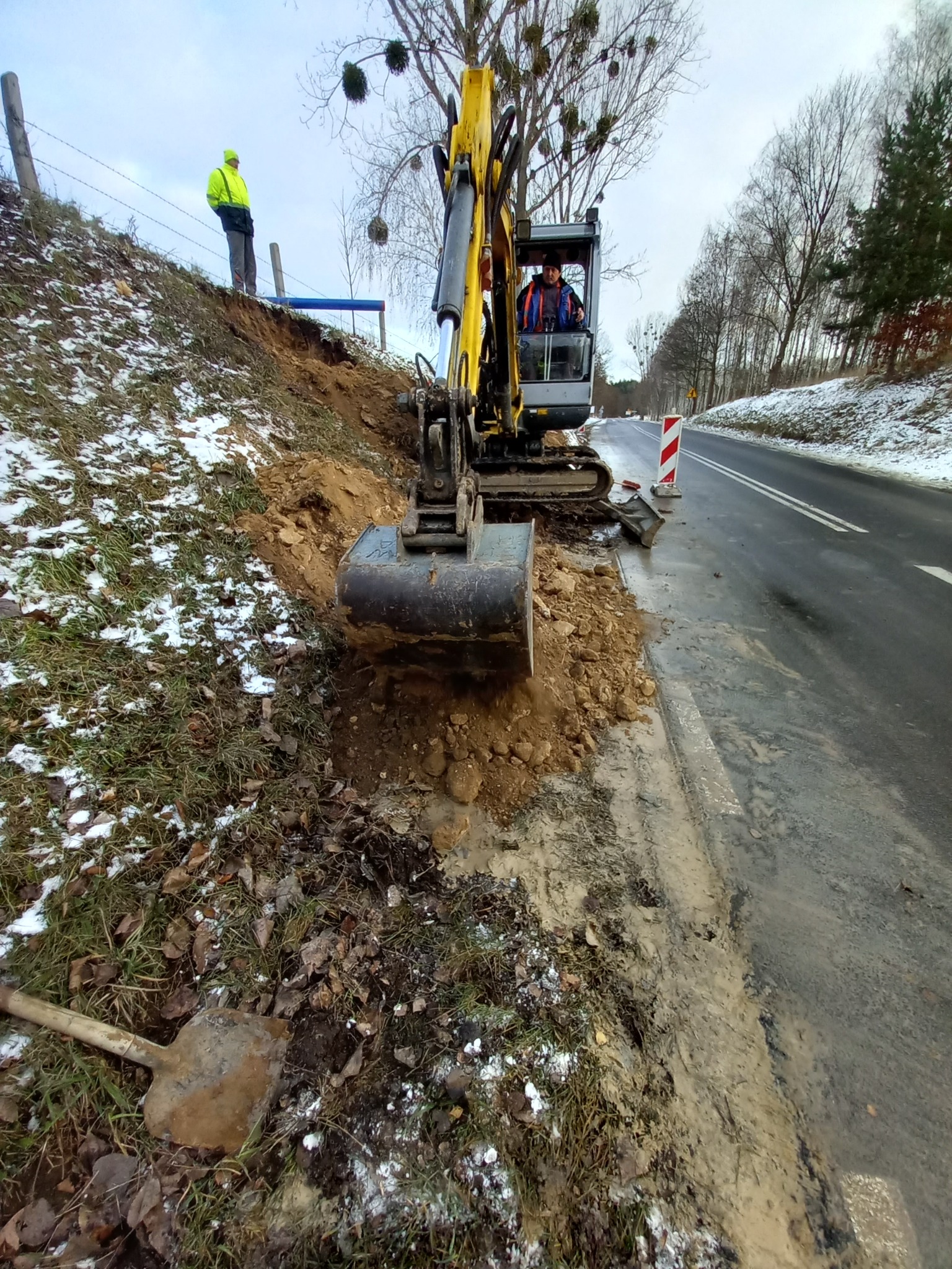 Żółta koparka przy drodze, operator w kabinie, obok pracownik w odblaskowej kamizelce, wykopany rów w ziemi, resztki śniegu na trawie, łopata leżąca na ziemi.