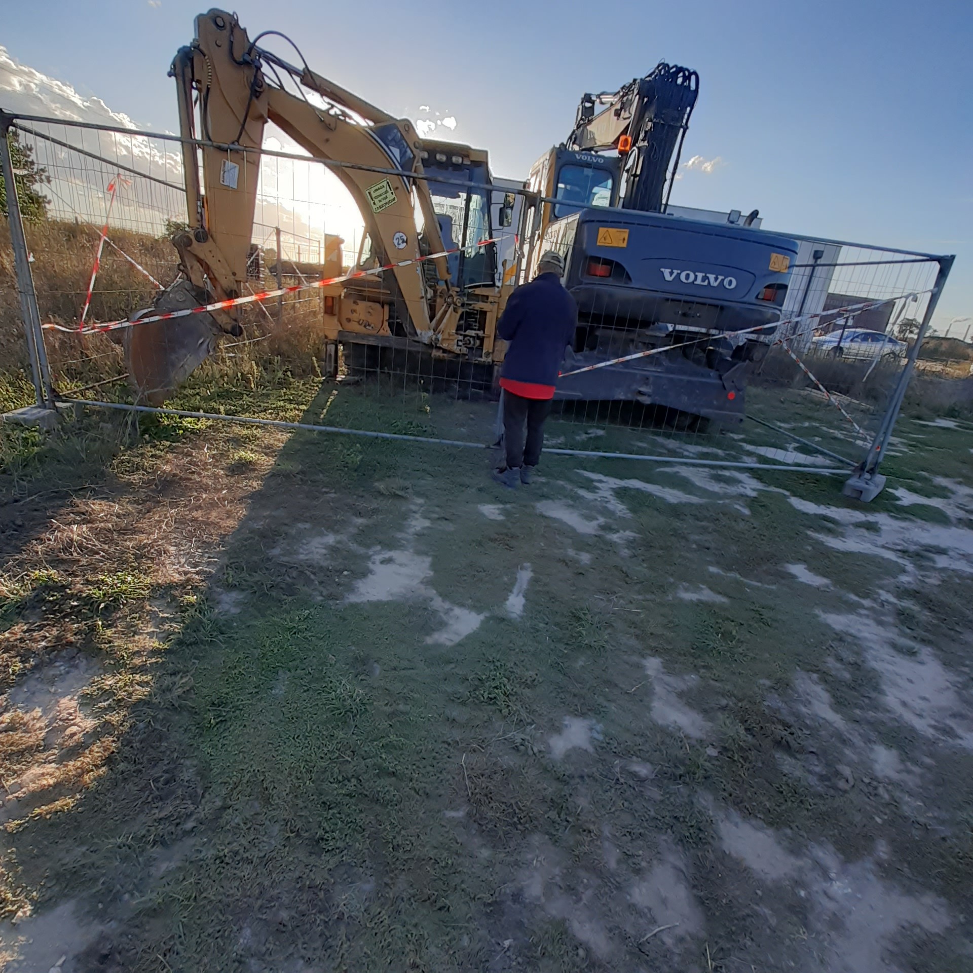 Dwie koparki, żółta i niebieska Volvo, stoją za ogrodzeniem z taśmą ostrzegawczą; osoba w kurtce stoi przed nimi na placu budowy z błotnistym podłożem.