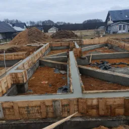 Wylane betonowe fundamenty domu jednorodzinnego z drewnianym szalunkiem, widoczne zbrojenia i hałda ziemi w tle.