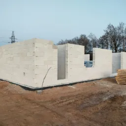 Wznoszone ściany domu z białych bloczków betonowych, widoczne palety z materiałem budowlanym, w tle słup wysokiego napięcia i drzewa.