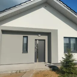 Nowoczesny dom jednorodzinny z elewacją w odcieniach szarości i bieli, widok na front z wejściem i oknami, małe drzewka iglaste przed budynkiem, częściowo zachmurzone niebo w tle.