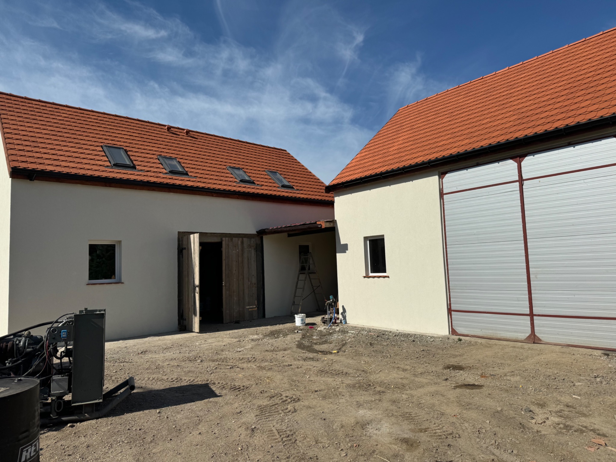Jasna elewacja budynku gospodarczego z czerwoną dachówką po tynkowaniu, widoczne otwarte drewniane wrota, okno, drabina oraz sprzęt malarski.