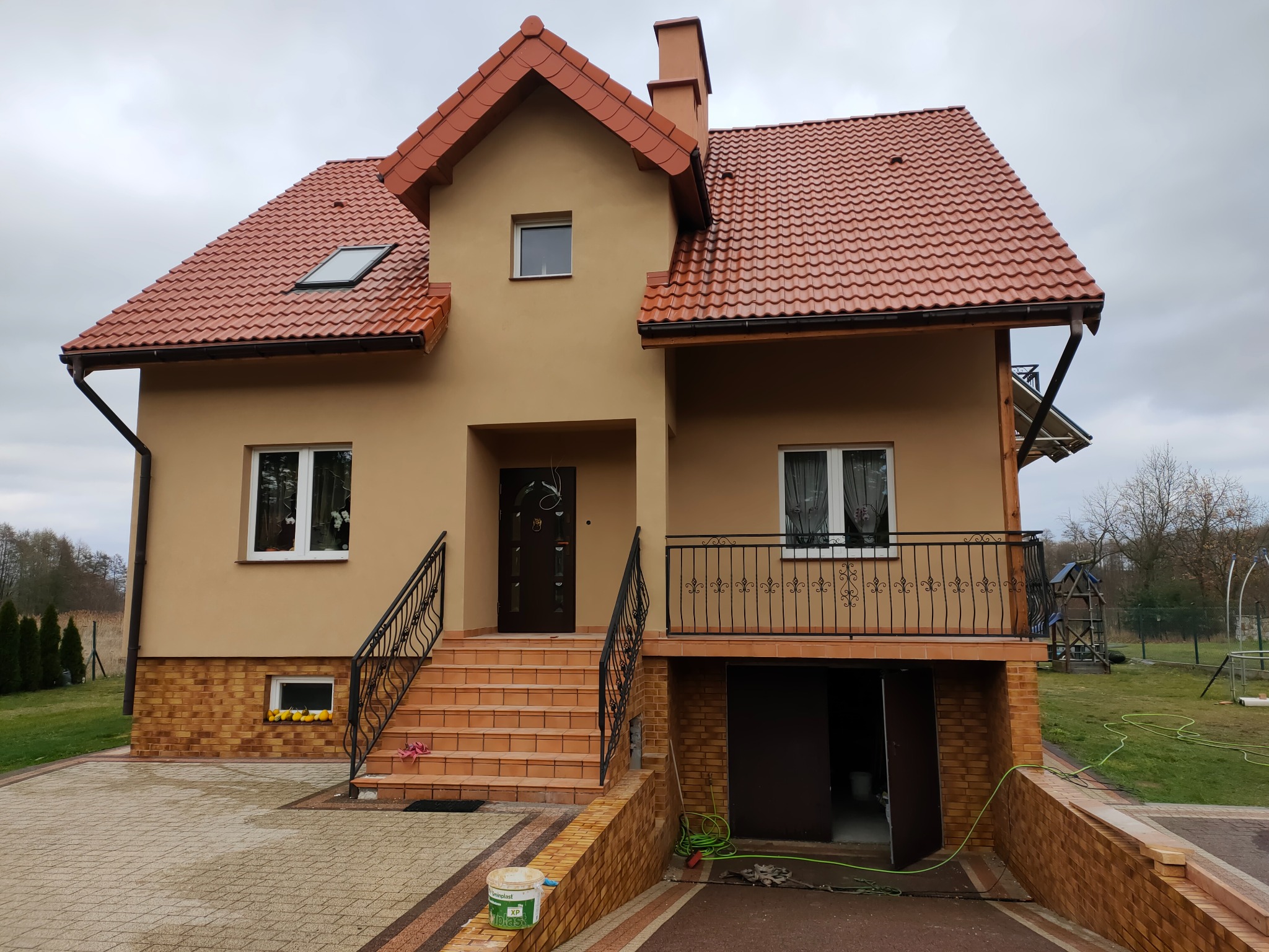 Dwupiętrowy dom z beżową elewacją i czerwoną dachówką, widoczne schody wejściowe z kutą balustradą, okna z białymi ramami i balkon z ozdobną barierką, boczna elewacja z wejściem do garażu.