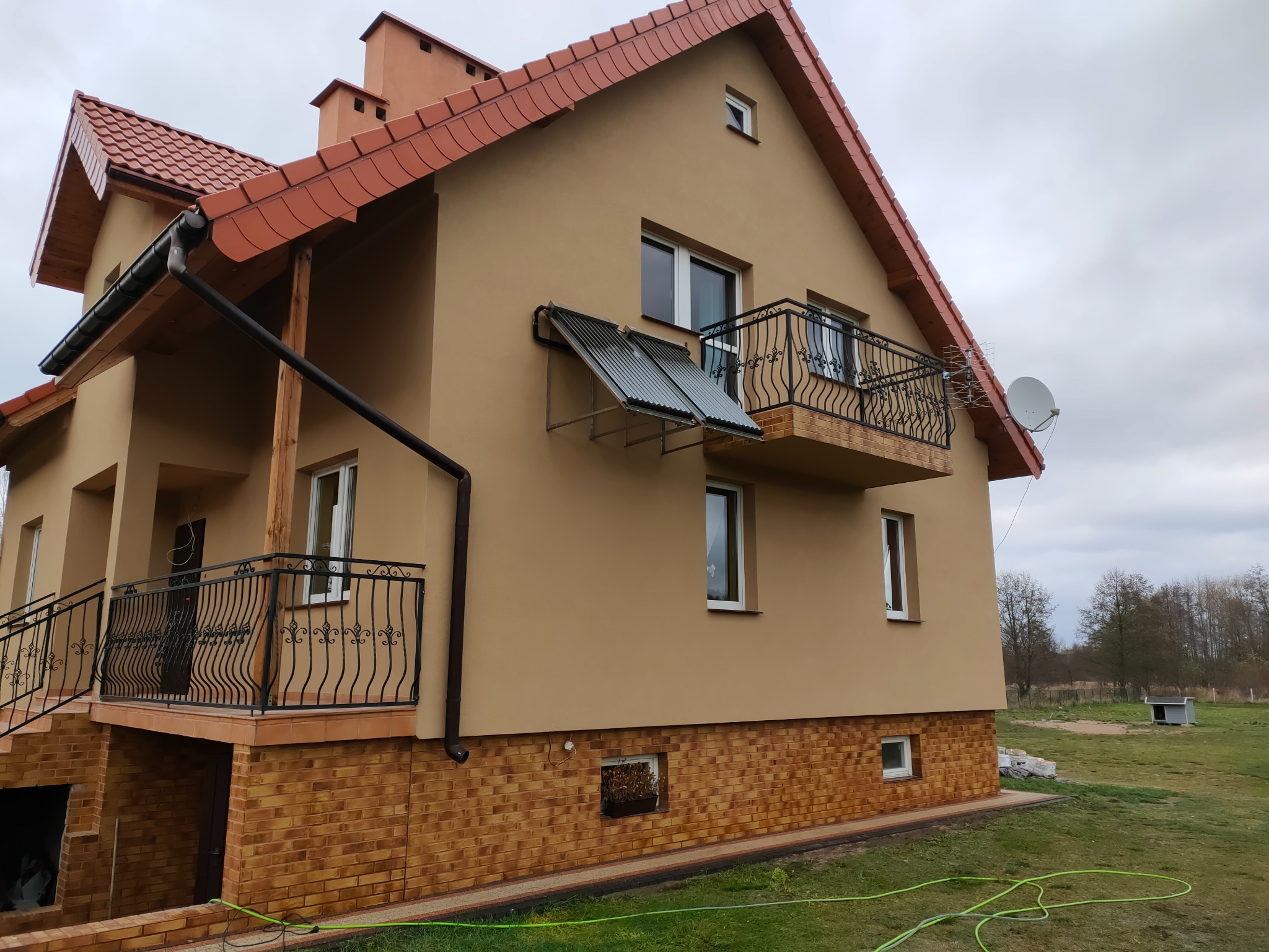 Dwupiętrowy dom z beżową elewacją, czerwoną dachówką, balkonami z kutą balustradą i kolektorami słonecznymi na ścianie, otoczony zielonym trawnikiem.