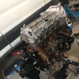 Terle-Garage - Silnik samochodowy na stojaku warsztatowym, górna część odnowiona i pomalowana na srebrno, dolna część brudna, widoczne ślady oleju, w tle biały samochód i narzędzia warsztatowe.