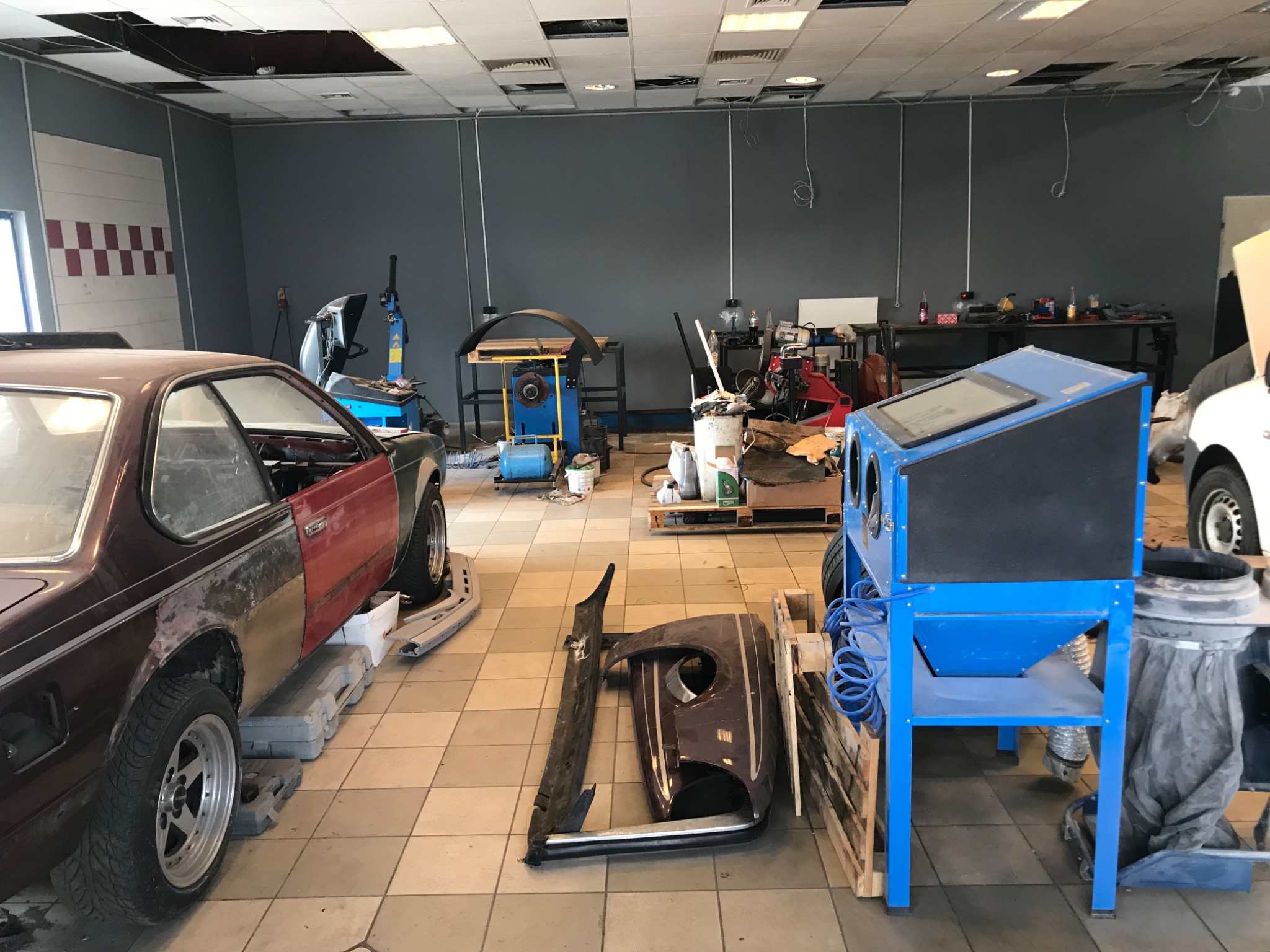 Warsztat samochodowy: brązowe BMW E24 na podnośniku, obok zdemontowany błotnik, w tle piaskarka i inne urządzenia warsztatowe.