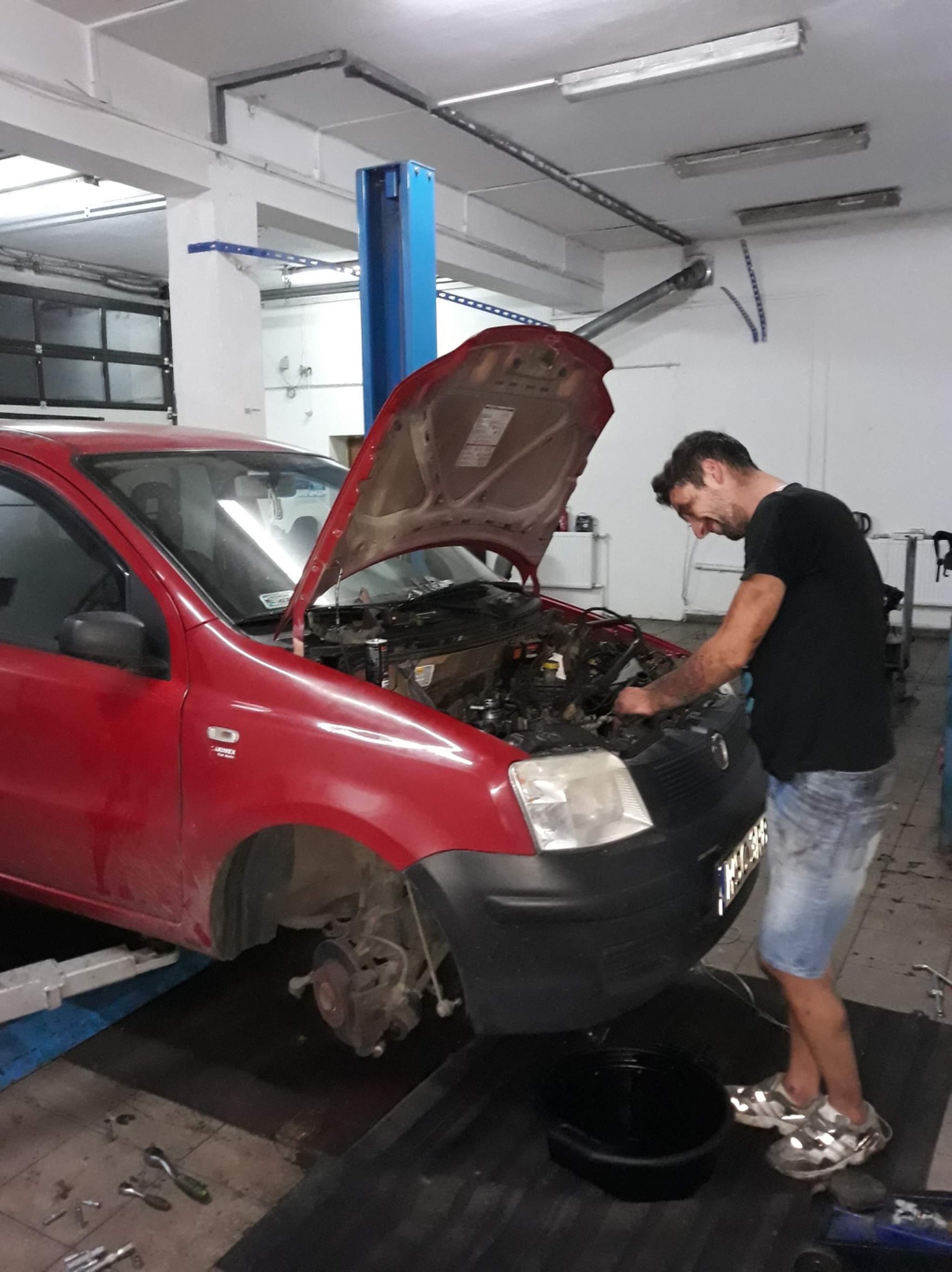 Czerwony samochód na podnośniku w warsztacie, z otwartą maską silnika, mechanik w dżinsowych szortach pochyla się nad silnikiem, obok miska na olej, narzędzia leżą na podłodze.