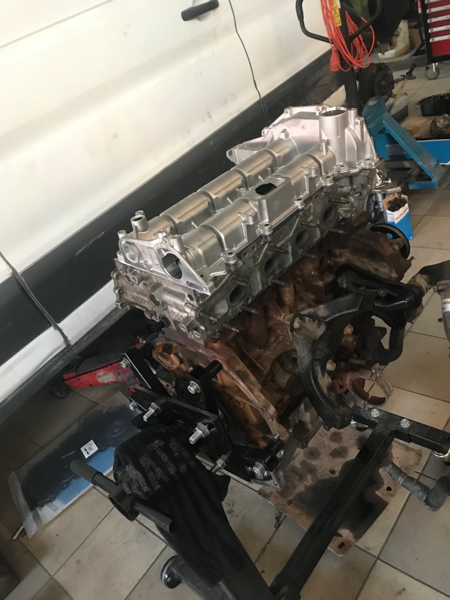 Silnik samochodowy na stojaku warsztatowym, górna część odnowiona i pomalowana na srebrno, dolna część brudna, widoczne ślady oleju, w tle biały samochód i narzędzia warsztatowe.