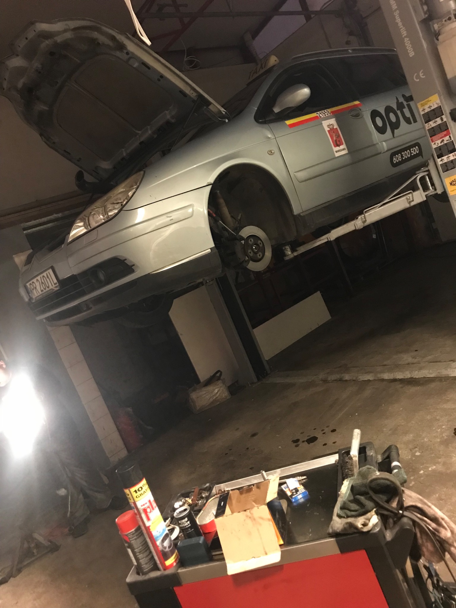 Samochód marki Citroen na podnośniku w warsztacie, z otwartą maską silnika i zdjętym kołem, widoczna tarcza hamulcowa. W tle narzędzia i akcesoria warsztatowe.