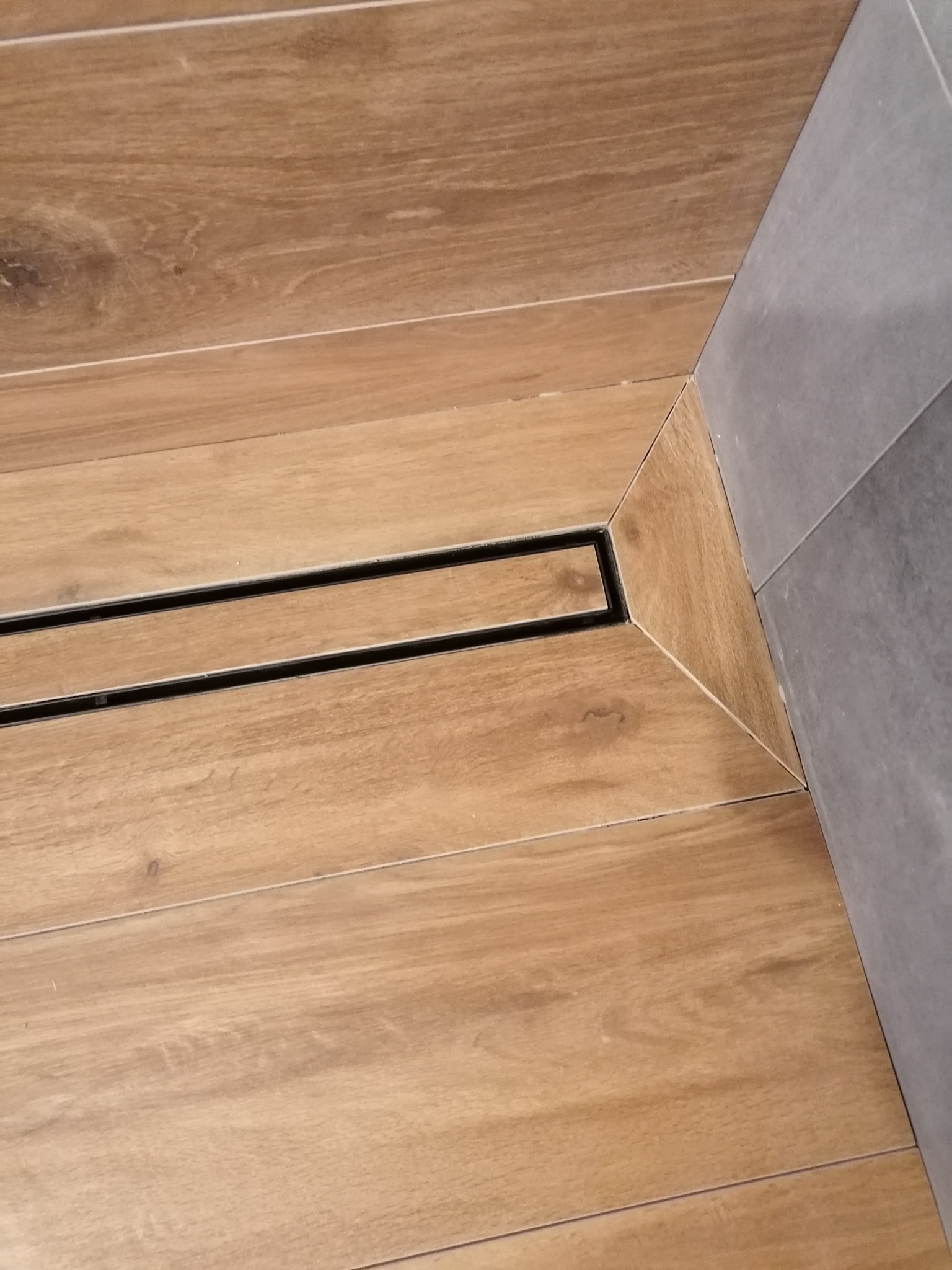 Szczegółowe ujęcie narożnika kabiny prysznicowej z odpływem liniowym i wykończeniem z płytek imitujących drewno oraz szarych płytek ściennych.