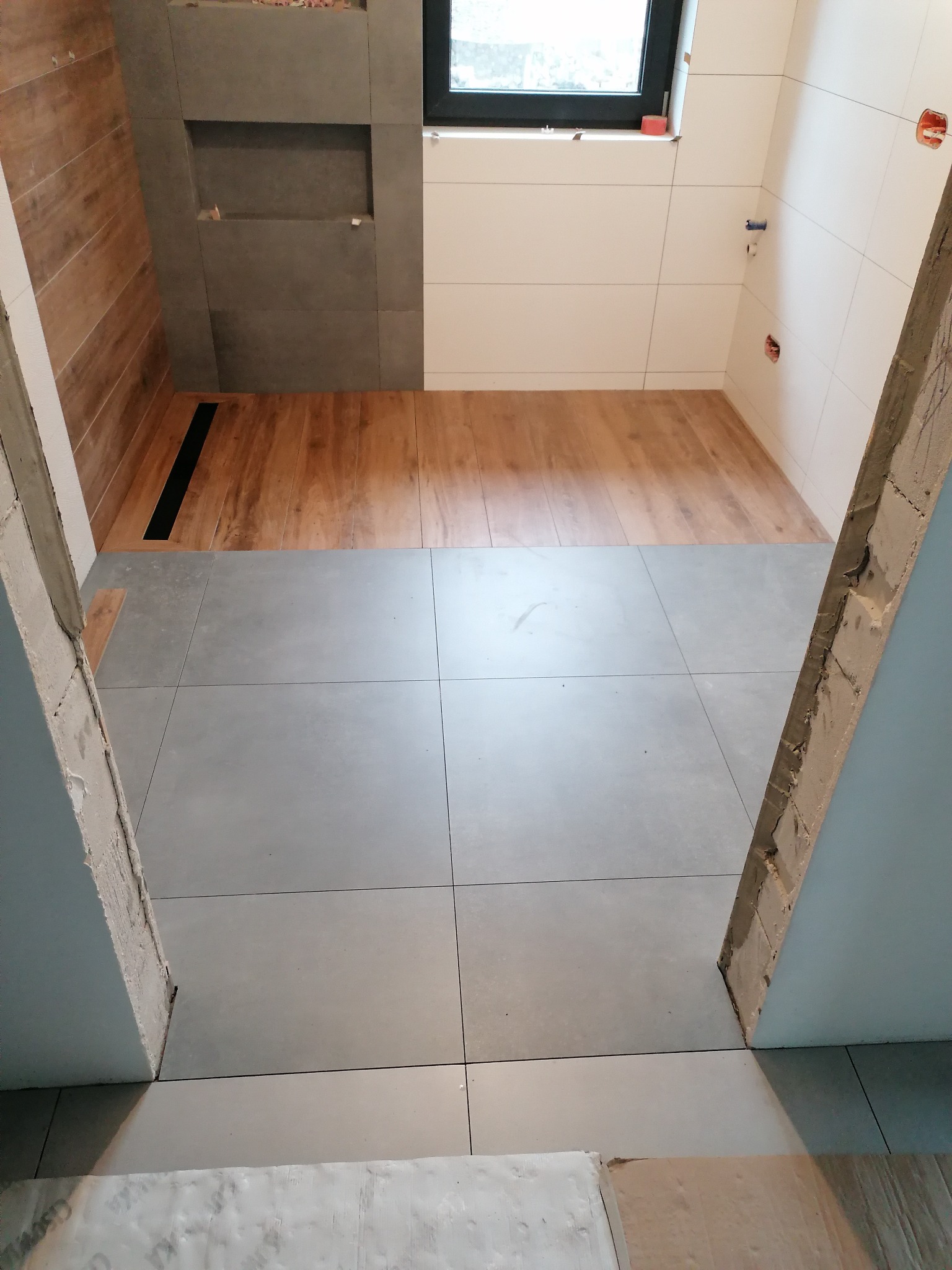 Łazienka w trakcie wykańczania: szare płytki podłogowe, drewnopodobne płytki w strefie prysznica z odpływem liniowym, białe płytki na ścianach, widoczne surowe futryny drzwi.