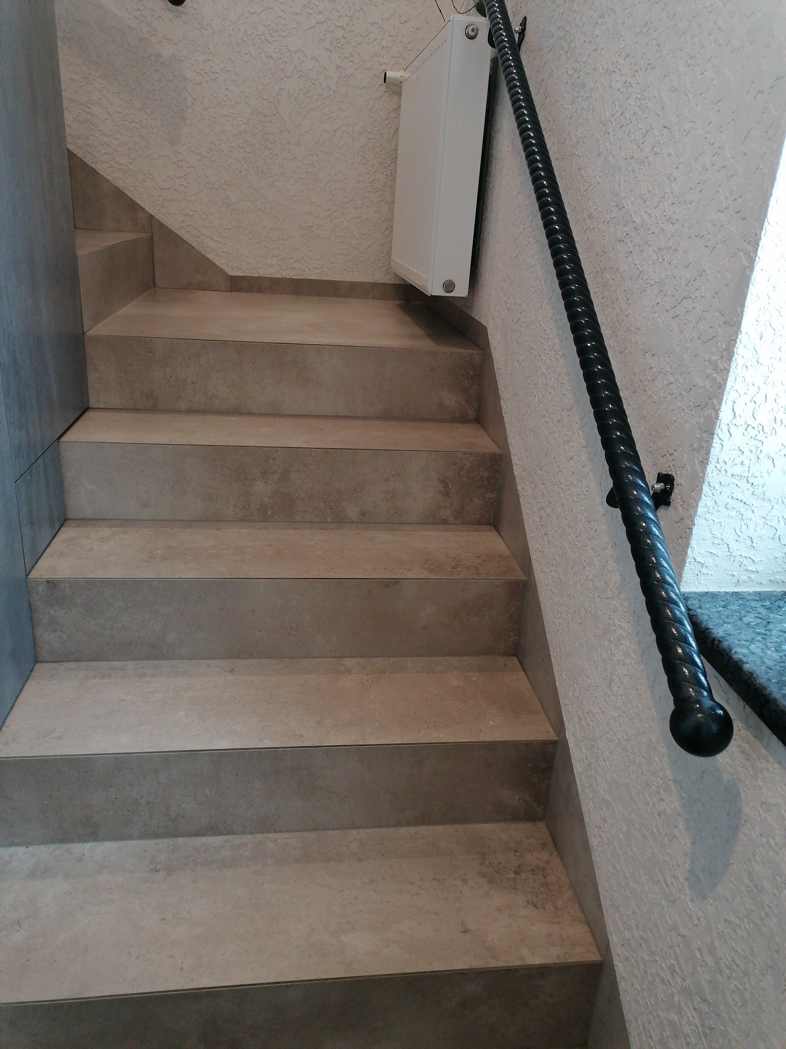 Schody wyłożone płytkami w odcieniach beżu, z ciemną, spiralną poręczą zamontowaną na teksturowanej ścianie. Biały grzejnik zawieszony nad schodami.