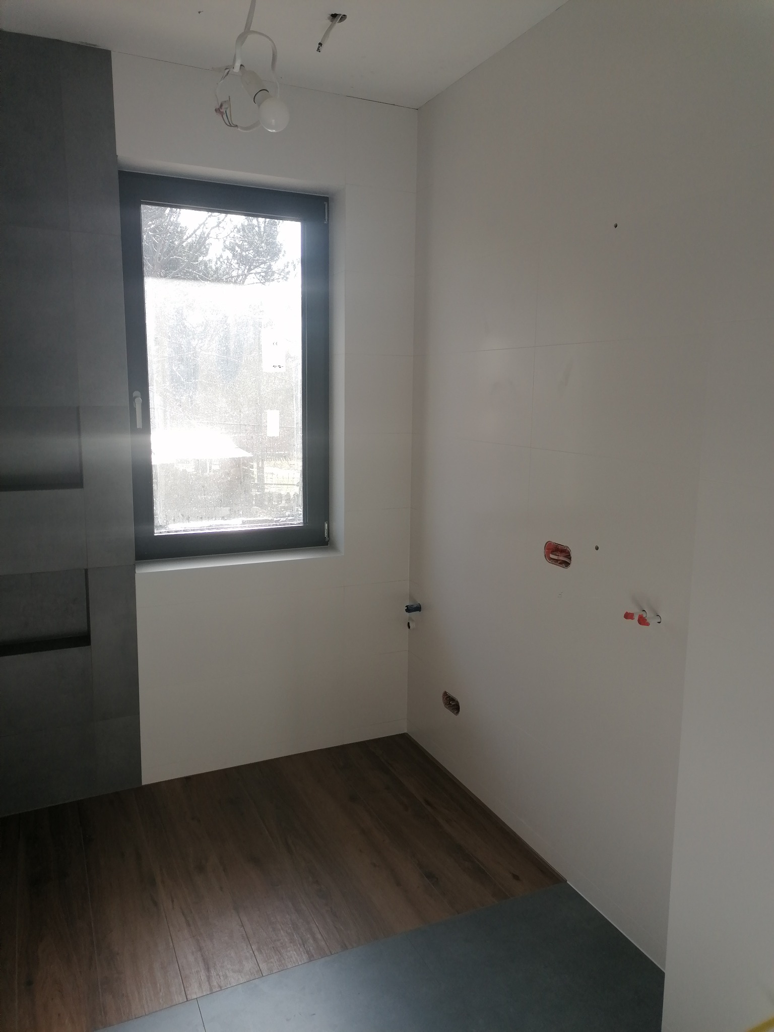 Wnętrze łazienki w trakcie remontu: częściowo położone płytki podłogowe imitujące drewno, szare płytki ścienne z wnękami, białe ściany z widocznymi instalacjami elektrycznymi i hydraulicznymi...