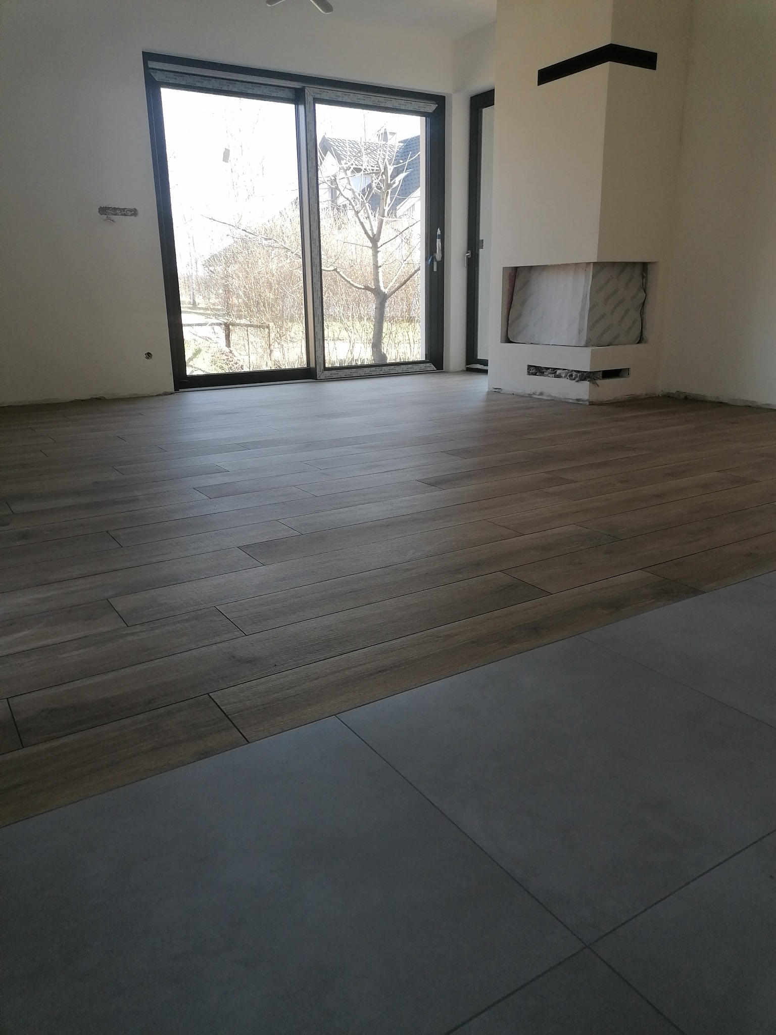 Układanie podłogi z płytek imitujących drewno w jasnym wnętrzu, widoczny fragment szarej podłogi z płytek, kominek w trakcie zabudowy i przesuwne drzwi balkonowe.