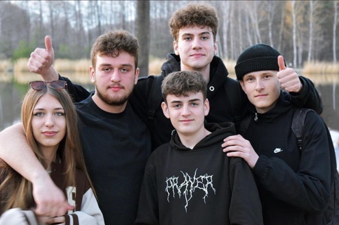 Grupa pięciu młodych osób pozuje do zdjęcia na tle drzew i wody, część z nich pokazuje kciuki w górę.