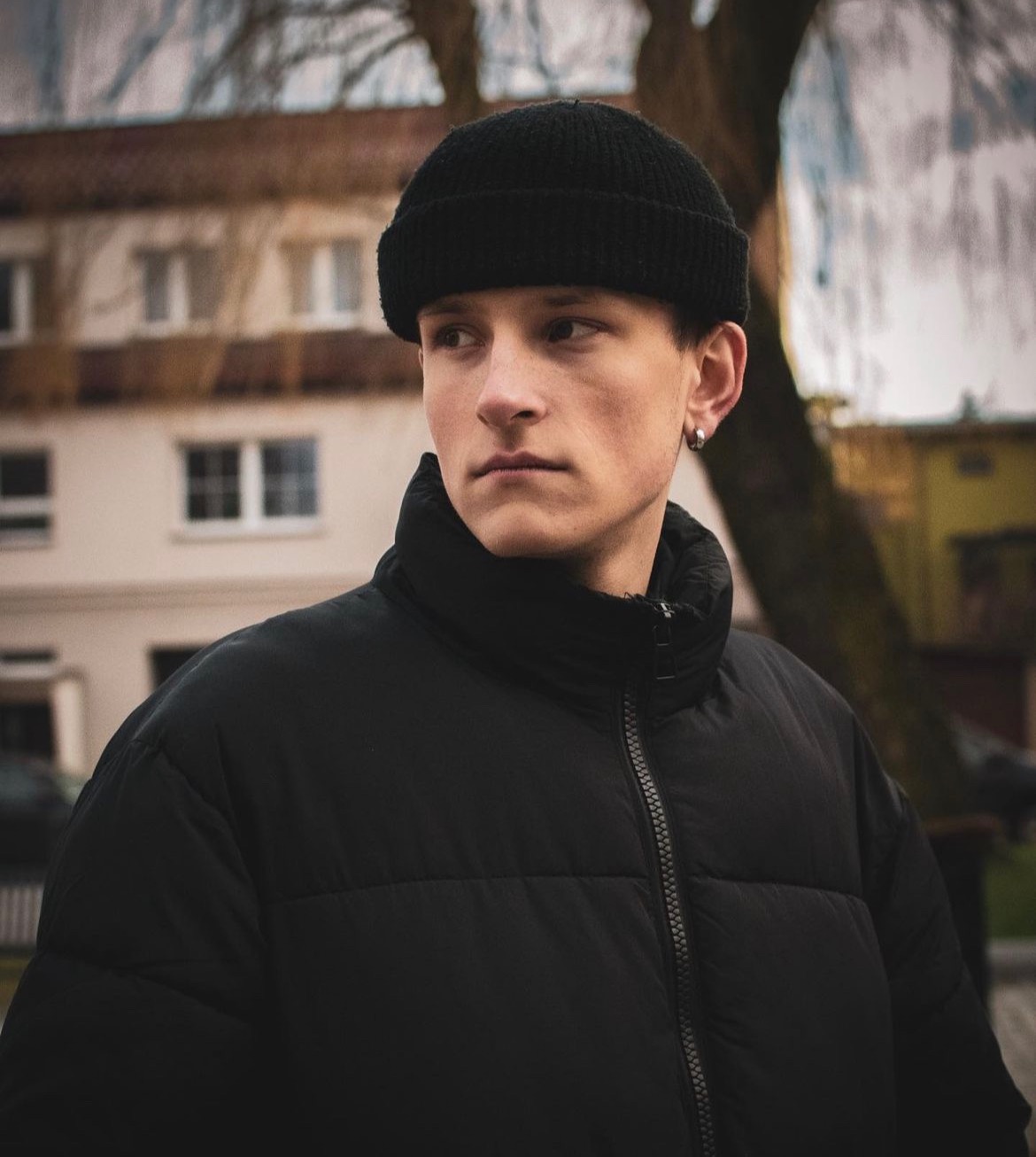 Portret młodego mężczyzny w czarnej kurtce i czapce, z kolczykiem w uchu, spoglądającego w lewo na tle budynku.