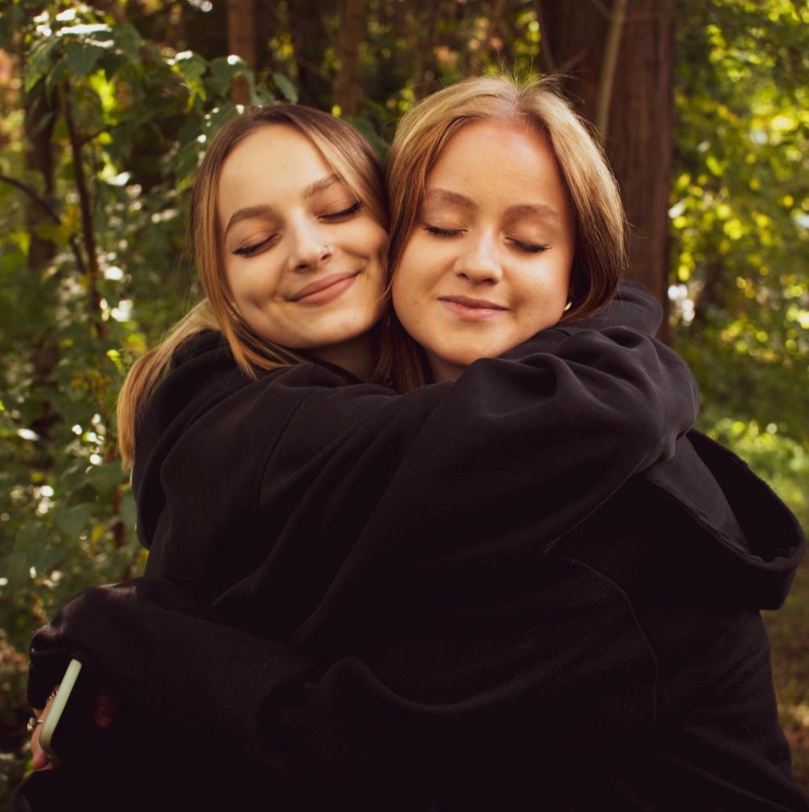 Dwie młode kobiety w czarnych bluzach z kapturem, przytulone w plenerze z zaroślami w tle, obie z zamkniętymi oczami i uśmiechem na twarzy.