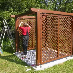 Montaż drewnianej altany z kratownicą: mężczyzna w czerwonej koszulce i czarnych ogrodniczkach przykręca elementy konstrukcji, obok drabina i narzędzia