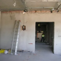 Wnętrze w trakcie remontu: ściany po nałożeniu gładzi, widoczne instalacje elektryczne, drabina oparta o ścianę, pracownicy na rusztowaniu w oddali.