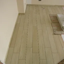 Ułożona podłoga z płytek imitujących drewno, widoczne kartony z opakowaniem płytek, ściana w jasnym kolorze, gniazdka elektryczne na ścianie.