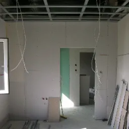 Wnętrze w trakcie prac z płytami kartonowo-gipsowymi, widoczne profile sufitowe, instalacje elektryczne wyprowadzone ze ściany, oraz otwarte okno w jasnym pomieszczeniu.