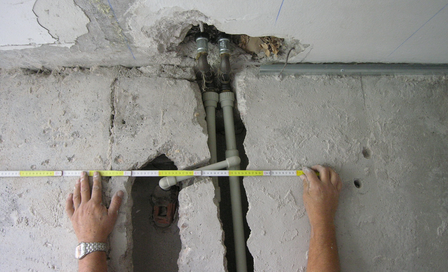Pomiar rur instalacji wodnej w ścianie podczas prac remontowych; widoczne odsłonięte rury miedziane i plastikowe, surowy beton i fragment sufitu z uszkodzeniami, a także ręce z miarką zwijaną.