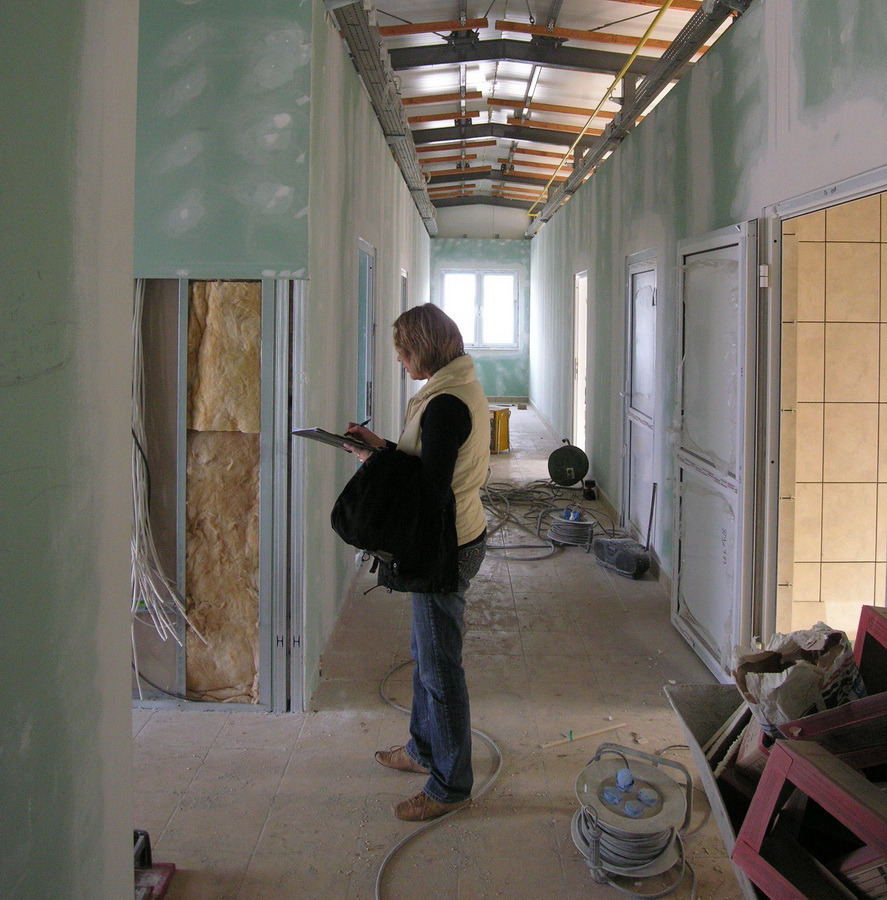 Korytarz w trakcie prac wykończeniowych: ściany z surową gładzią, widoczna izolacja w otworze drzwiowym, kobieta z notatnikiem sporządza uwagi, na podłodze zwinięte kable i resztki materiałów...