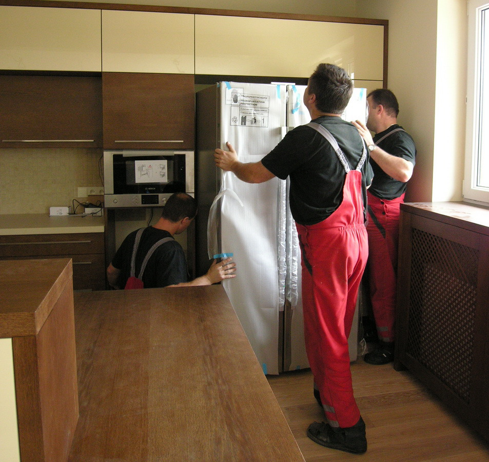 Trzech monterów w czerwonych kombinezonach instaluje nową lodówkę w kuchni z zabudową w odcieniach brązu i beżu, lodówka w folii ochronnej, widoczny mikrofalówka Bosch i grzejnik.