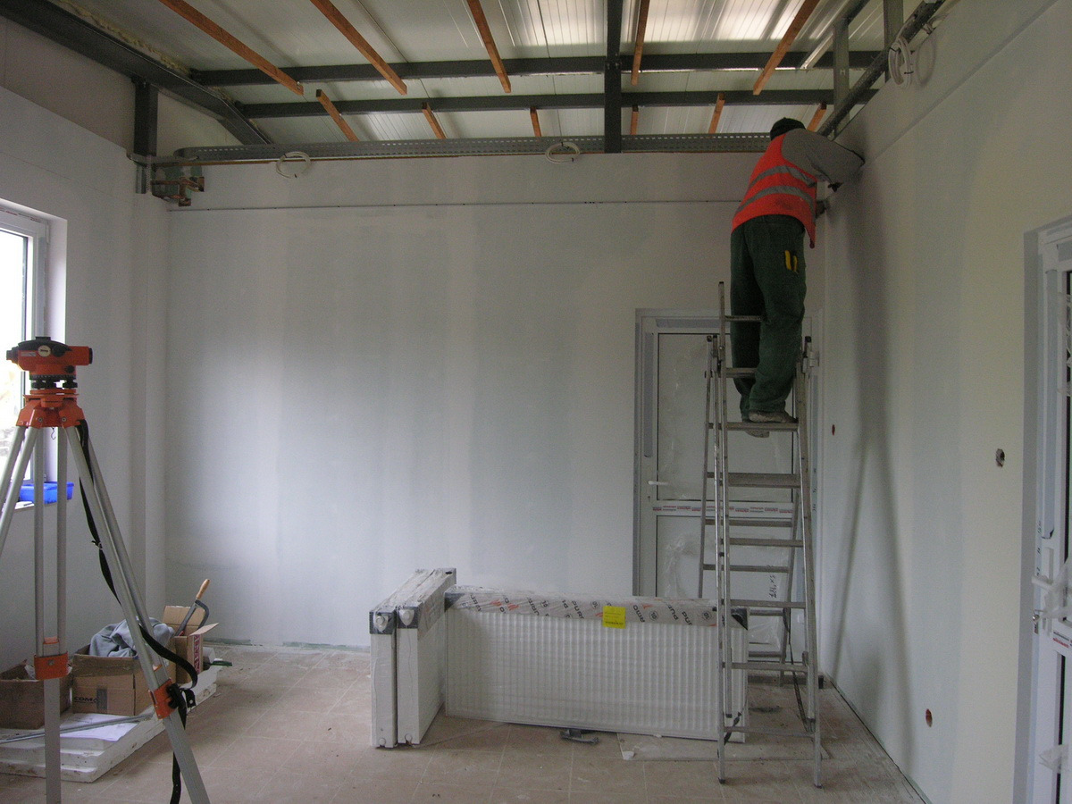 Pracownik w kamizelce odblaskowej na drabinie mocuje elementy do sufitu w pomieszczeniu z wygładzonymi ścianami, widoczny niwelator optyczny na statywie.