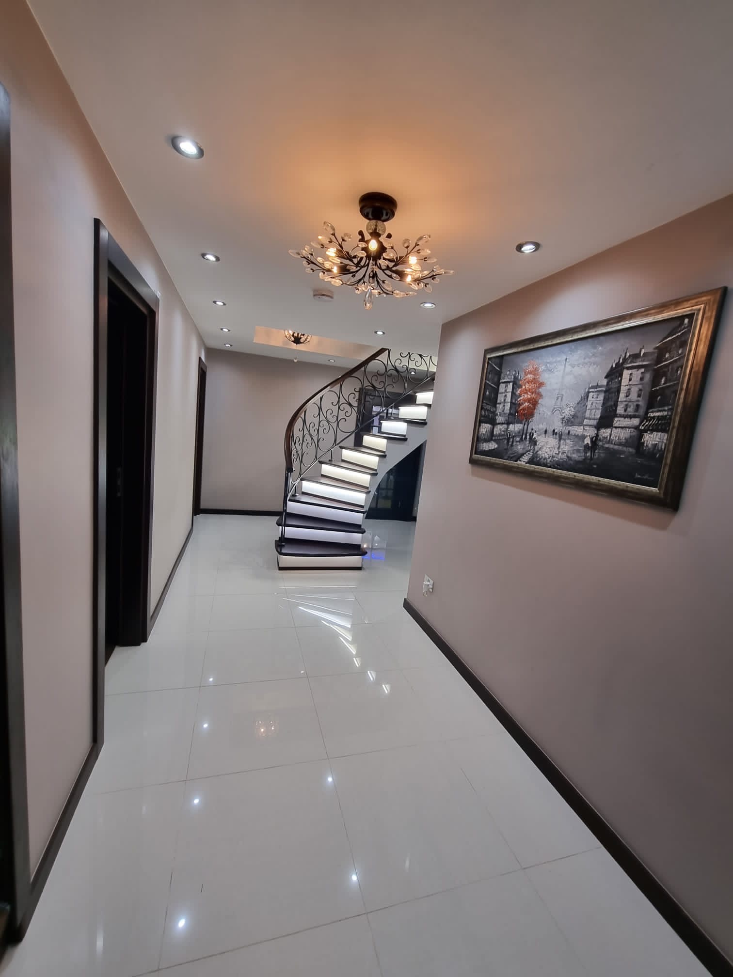 Elegancki korytarz z krętymi schodami z podświetleniem LED, ozdobnym żyrandolem i obrazem z motywem miejskim na ścianie.