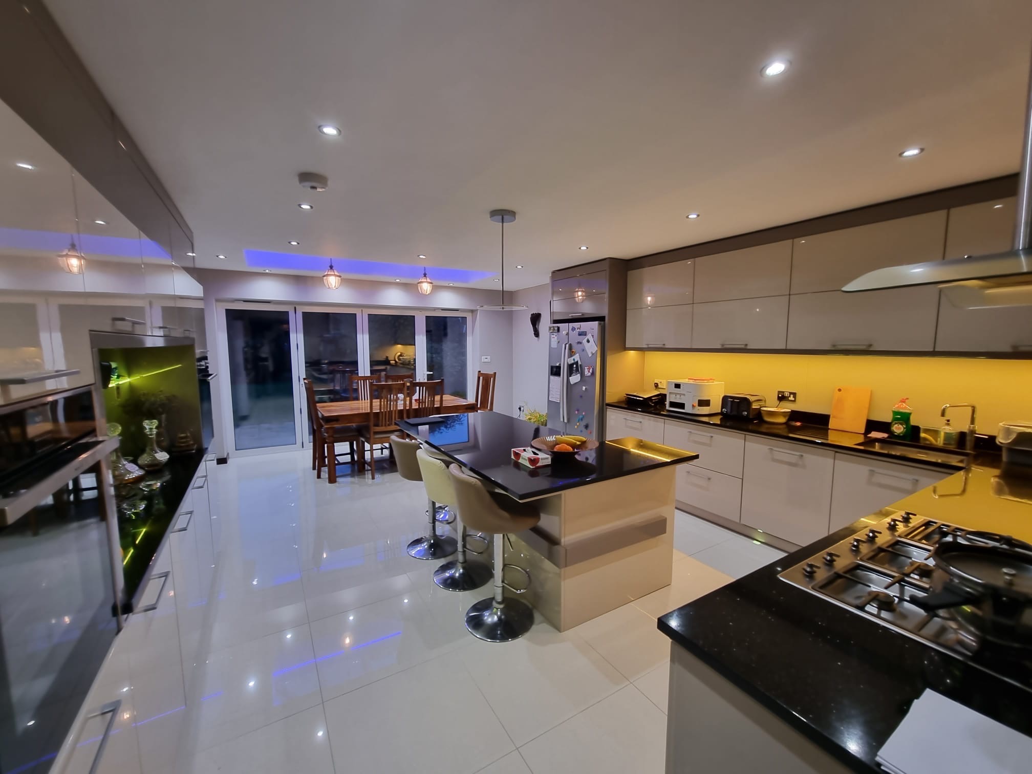 Przestronna, nowocześnie urządzona kuchnia z wyspą i barkiem, oświetlona punktowymi lampami i podświetleniem LED, widok na jadalnię i przesuwne drzwi balkonowe.