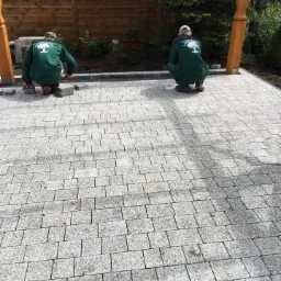 Dwóch pracowników w zielonych kombinezonach z logo drzewa układa szare kostki na nowo powstającym chodniku z kostki, widoczne drewniane elementy konstrukcji i ogrodzenie w tle.