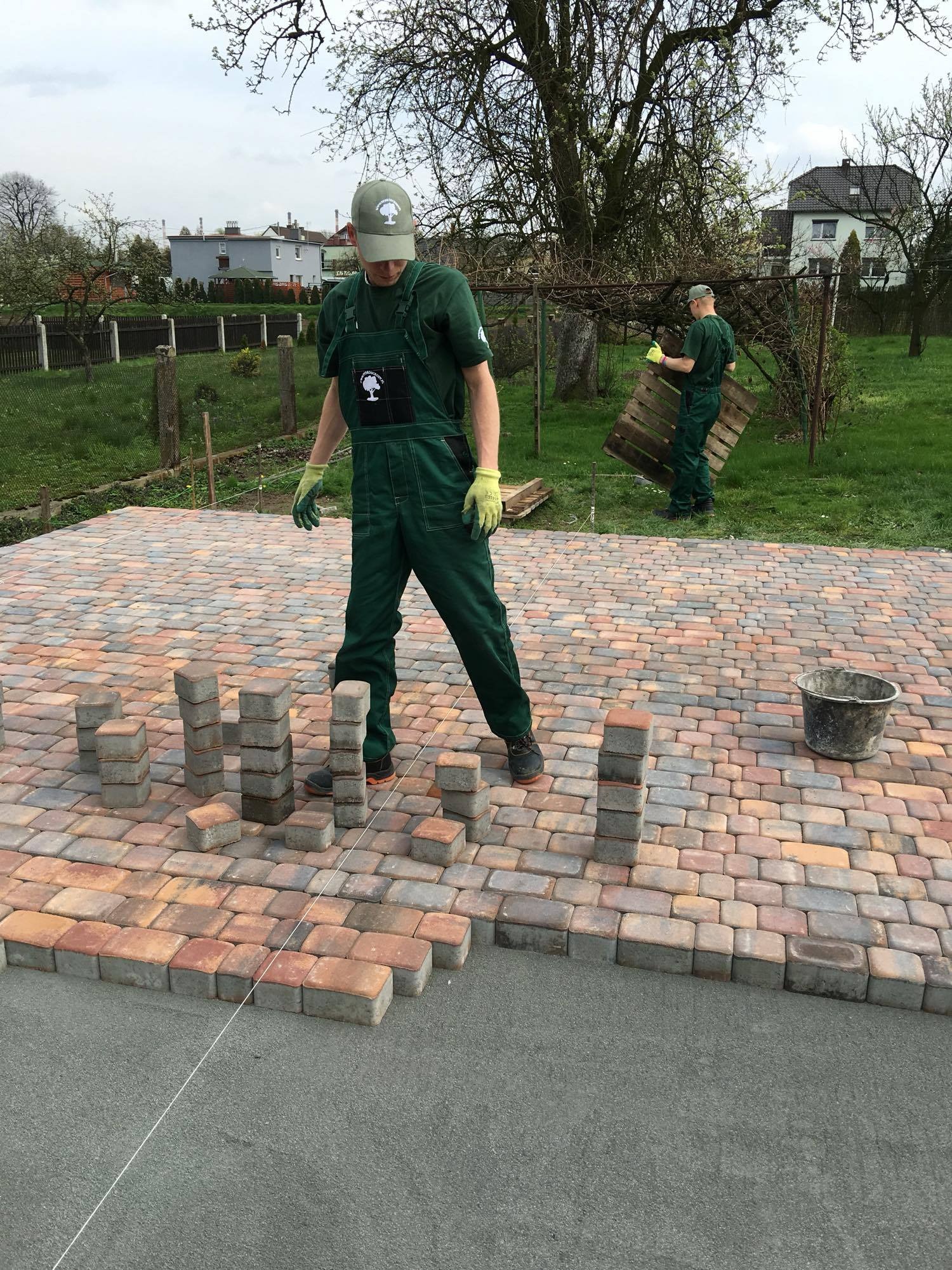 Pracownik w zielonym kombinezonie układa wielobarwną kostkę brukową, tworząc kolumny z pojedynczych elementów. W tle drugi pracownik z drewnianą paletą i widoczne zabudowania.