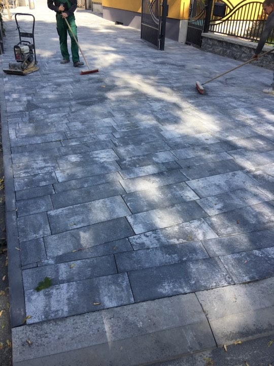 Świeżo ułożona, szara kostka brukowa w kształcie prostokątów, z widocznymi pracownikami wygładzającymi powierzchnię za pomocą zamiatacza i ubijarki.