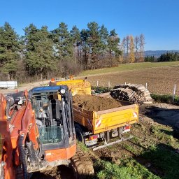 Firma Uslugowa Wojciech Łukaszka - Pomarańczowa koparka i żółta wywrotka wypełniona ziemią na tle pola i lasu w słoneczny dzień. Widoczne ślady pracy ziemnych.