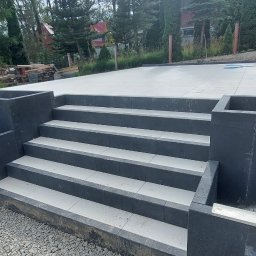 Firma Uslugowa Wojciech Łukaszka - Schody zewnętrzne z szarych płyt, wykończone ciemnym granitem, prowadzące do jasnego podjazdu. W tle drzewa iglaste i budynek z czerwonym dachem.