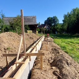 Firma Uslugowa Wojciech Łukaszka - Fundamenty w szalunku z drewnianymi podporami na tle wiejskiego krajobrazu pod błękitnym niebem. Widoczny pracownik w oddali.